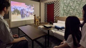 A hotel room with a bed, a desk, a projector screen displaying an image of animals, and a wall with a leaf pattern.