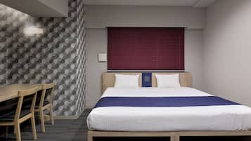 A hotel room with a bed, a desk, two chairs, a window with blinds, and a patterned wall.