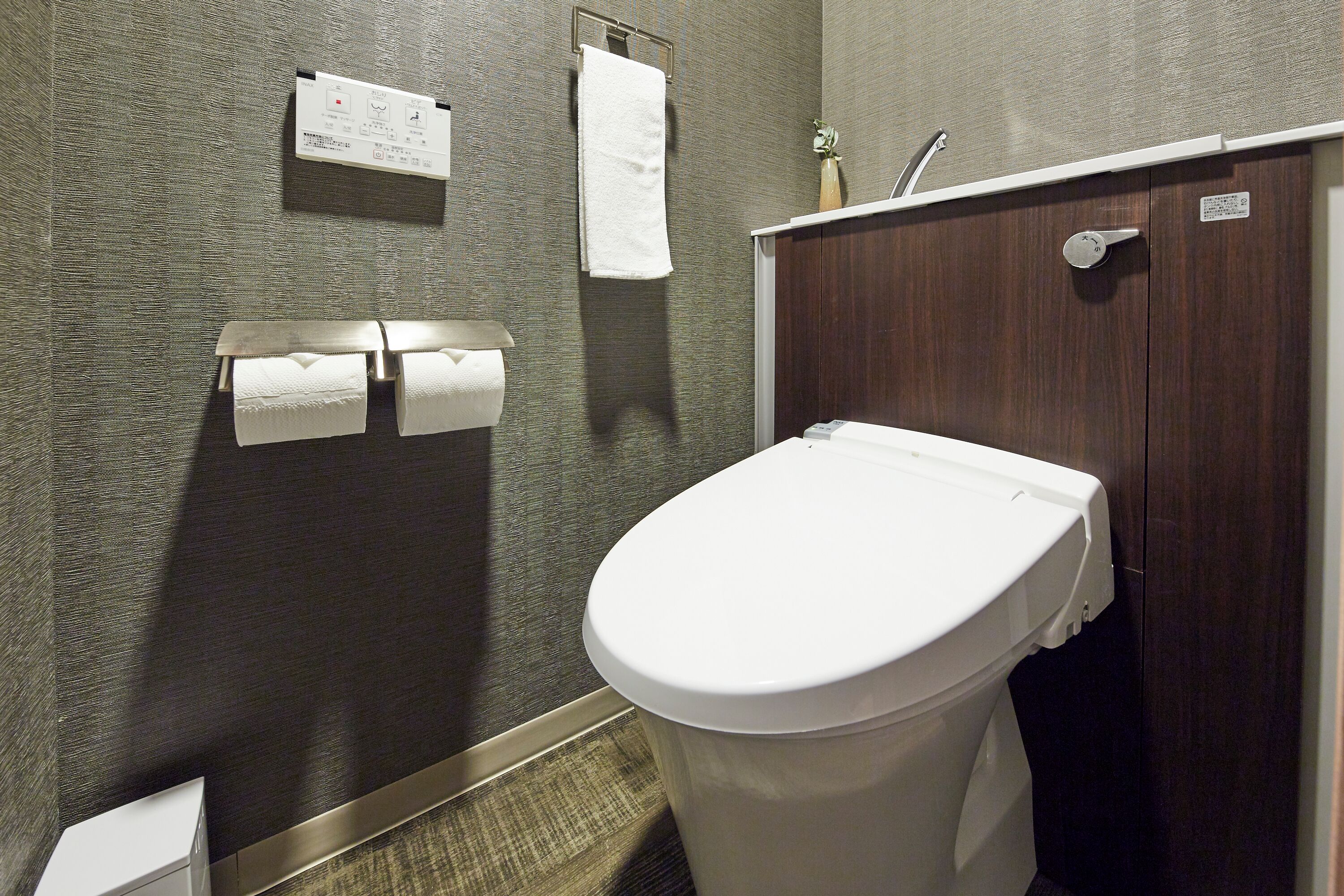 A bathroom with a toilet, a towel rack with towels, a paper towel dispenser, and a wall-mounted thermostat.
