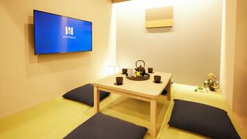 A room with tatami flooring, a low table with a tea set, and a flat-screen TV displaying "MONDAY".