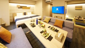 A hotel room with a bed, a sofa, a dining table set with a fruit bowl, a television, and a desk with a lamp. - MONday Apart Premium Hamamatsucho (Tokyo)