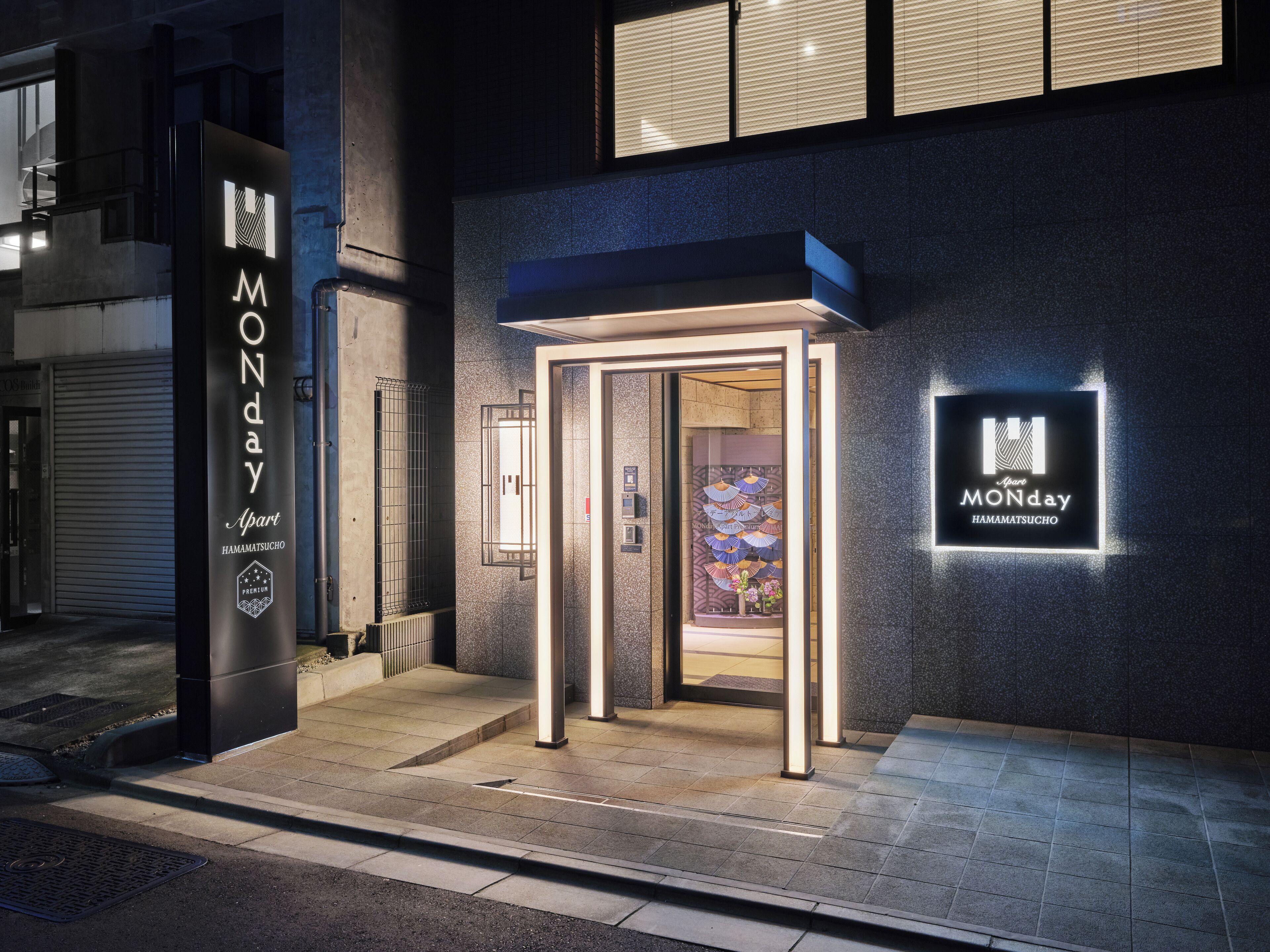 A hotel entrance with a modern design, a sign indicating "MONDAY HAMAMATSUCHO", and a glass door leading into the building.