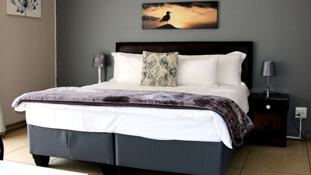 Una cama bien hecha con un cabecero oscuro, almohadas blancas y una manta decorativa estampada. Junto a la cama, una mesita de noche con una lámpara.