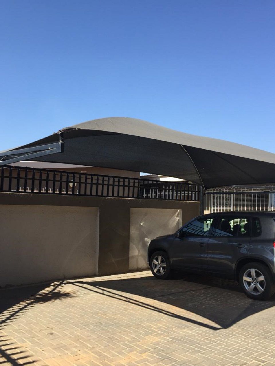 Un auto estacionado bajo una zona sombreada, con una cerca de metal y un edificio al fondo.