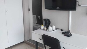 Una habitación pequeña y limpia con un escritorio blanco, una silla negra, un televisor de pantalla plana y una cafetera.