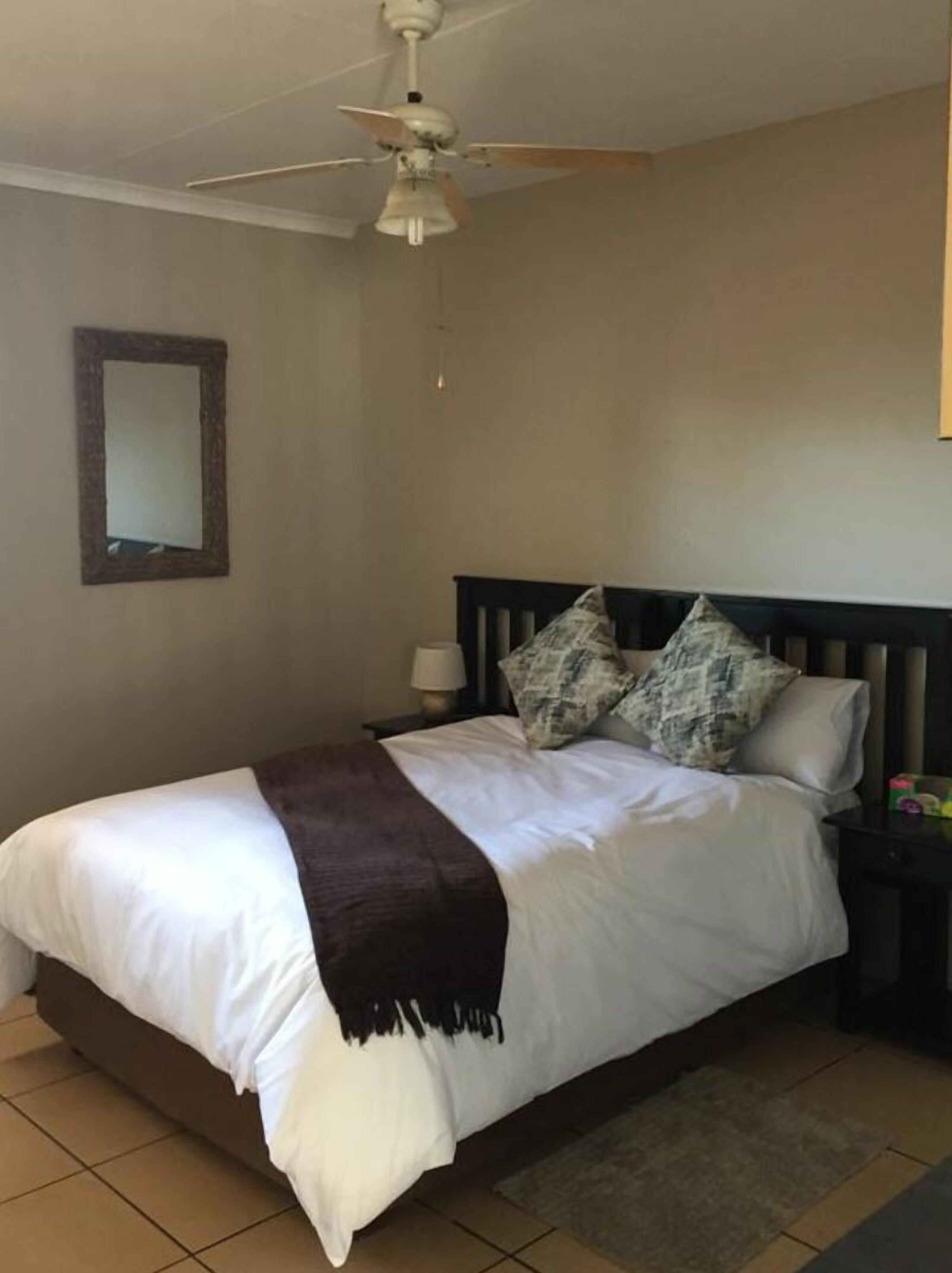 Een slaapkamer met een bed, nachtkastje, lamp, plafondventilator en een spiegel aan de muur.
