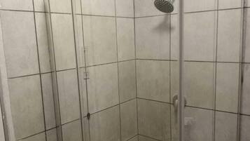 Un box de ducha con paredes de vidrio, piso a cuadros, ducha de mano y estante metálico.