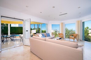 Ein großzügiges Wohnzimmer mit einer breiten beigen Couch, einem Essbereich mit Blick auf den Pool und großen Fenstern, die einen Ausblick ins Freie bieten.