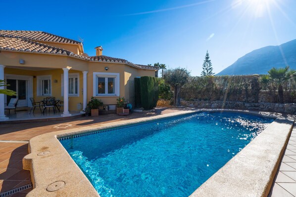 Ein Schwimmbad mit Fliesen umgeben, eine Terrasse mit Stühlen und Tischen sowie ein Berg im Hintergrund.