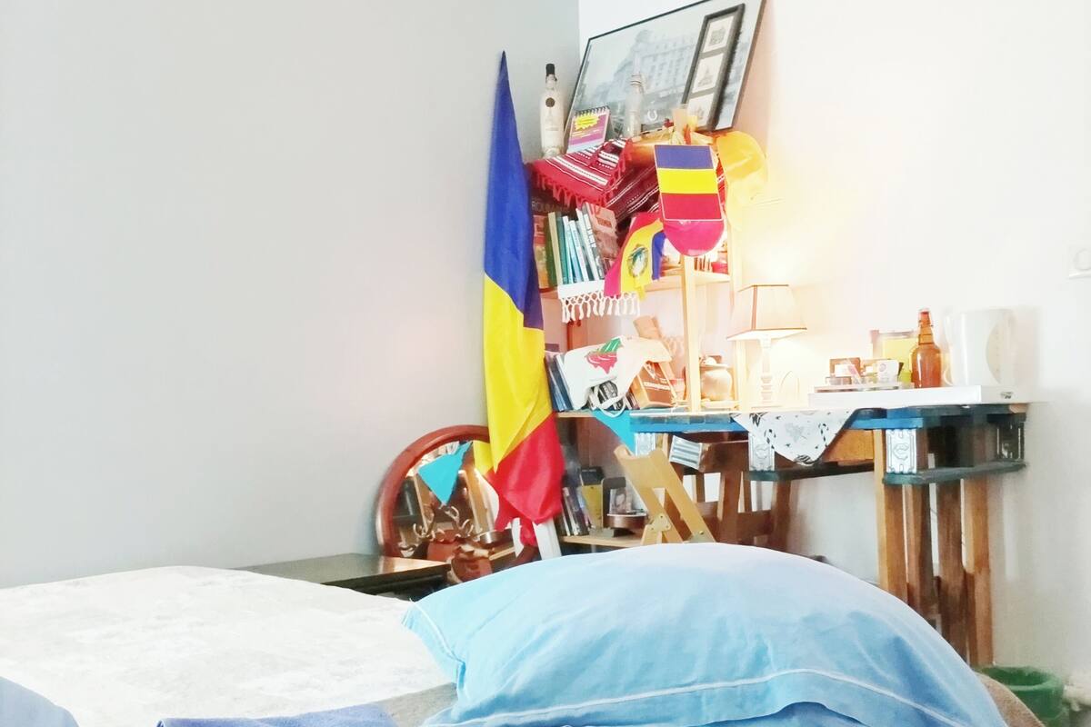 A bedroom with a bed, a desk, and a chair. There is a flag on the wall and a lamp on the desk.