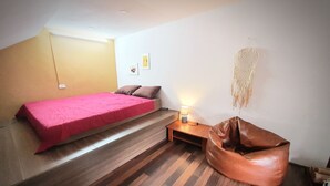 A bedroom with a bed, a wooden bench, a bean bag, and a wall-mounted macrame piece.