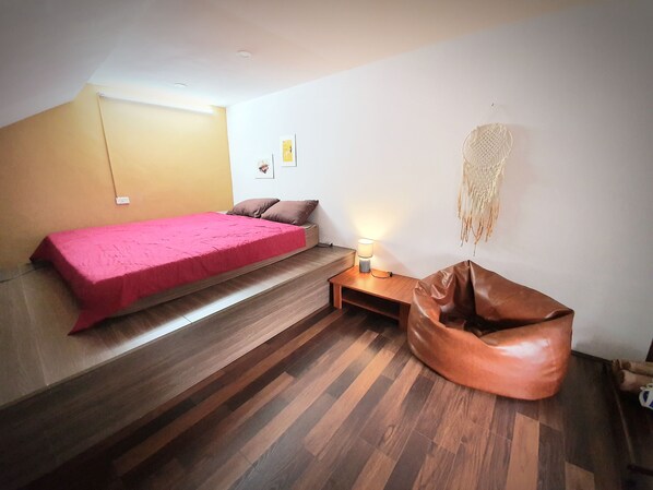 Un dormitorio con una cama, un banco de madera, un sillón de peluche y un macramé colgado en la pared.