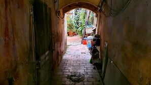 A narrow, brick-paved alleyway with a rustic, weathered appearance, leading to a small courtyard with potted plants and outdoor seating.