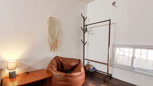 A room with a wooden floor, a brown bean bag chair, a small wooden table with a lamp, a wall-mounted macrame piece, a coat rack, and a window with blinds.