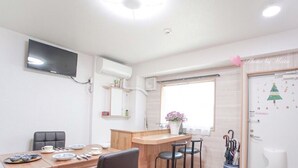 Ein moderner, hell erleuchteter Raum mit einem Holzespeisetisch, Stühlen, einem an der Wand befestigten Fernseher, einem Fenster mit Jalousien und einer Tür mit weihnachtlicher Dekoration.