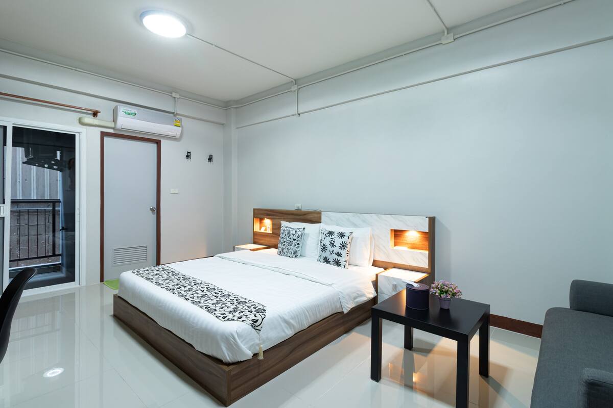 A modern hotel room with a large bed, bedside tables, a small table with a plant, and a window with a view.