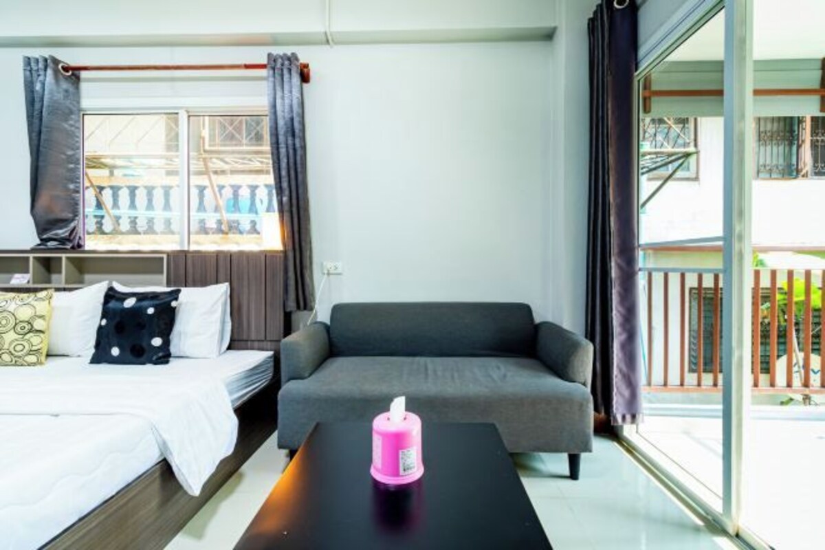 A modern room with a bed, a grey sofa, a black table with a pink object, and a large window with curtains.