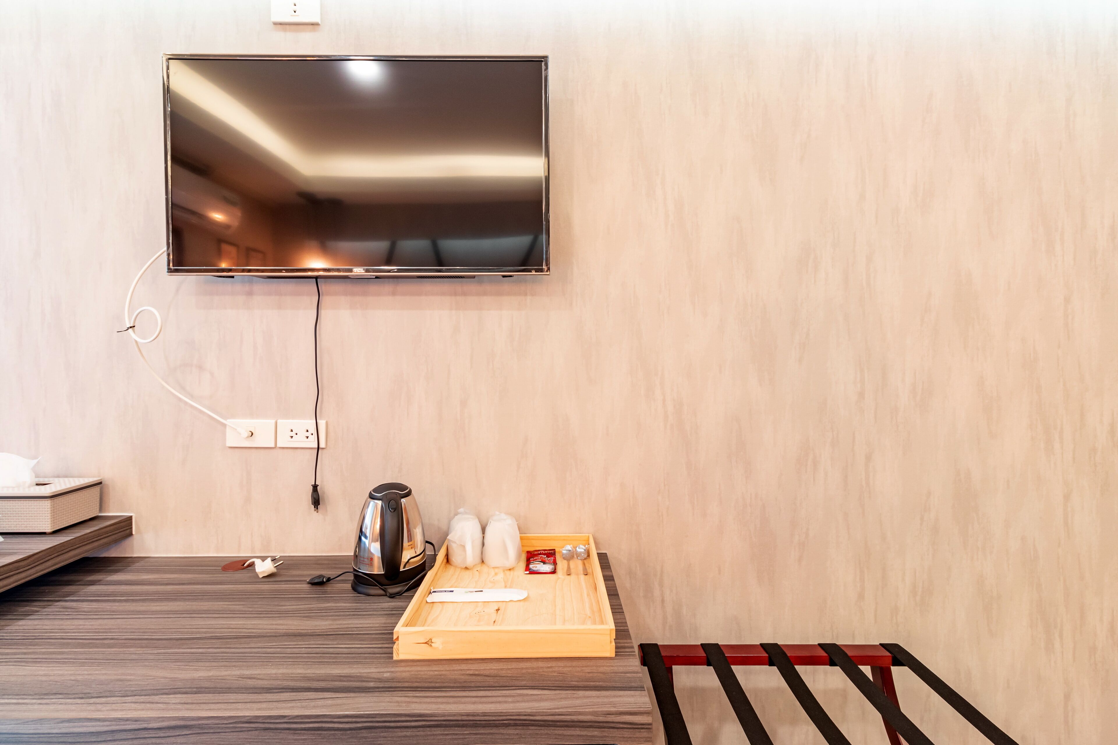 A hotel room with a flat-screen TV mounted on the wall, a wooden tray with tea bags and a kettle, and a red bench.
