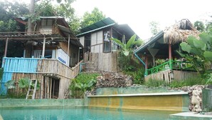 A pool area with a wooden deck, a stone wall, and a thatched-roof structure. - Baansuan Chomchun (Mae Taeng)