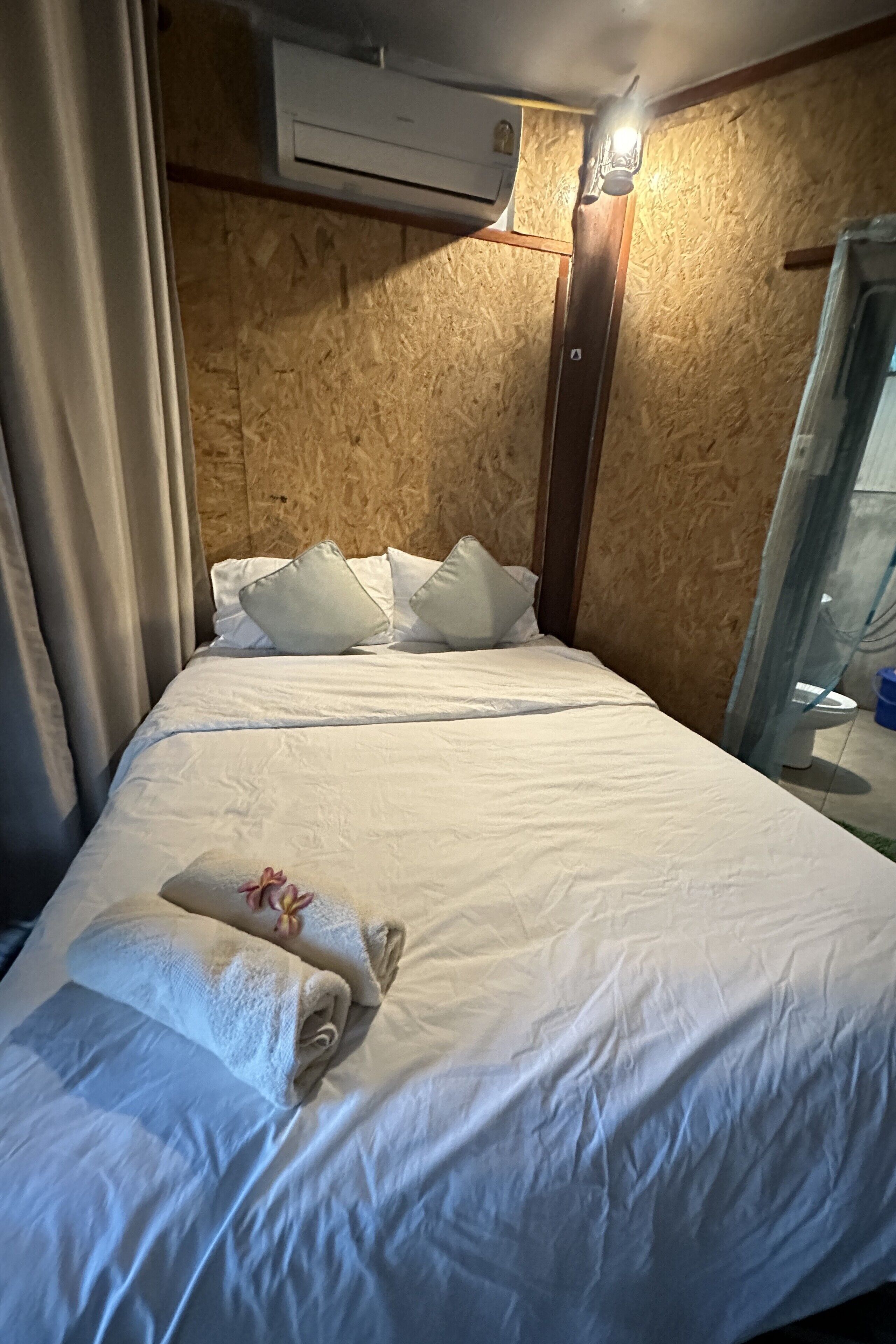 A neatly made bed with white linens and pillows, an air conditioning unit above, and a small bedside table with a lamp.