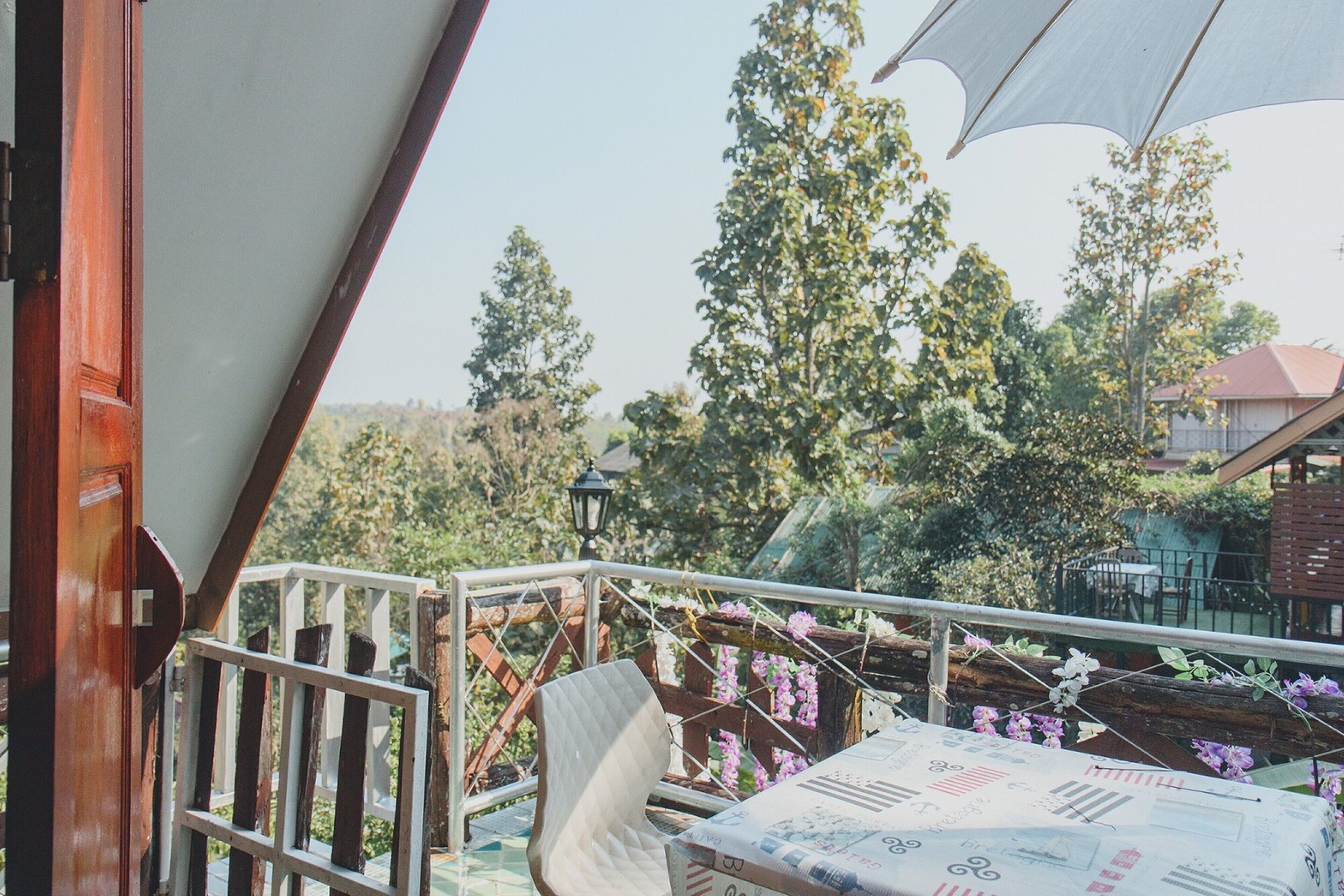 Un balcon avec une chaise blanche, une table recouverte d’une nappe à motifs et une vue sur les arbres et les bâtiments.