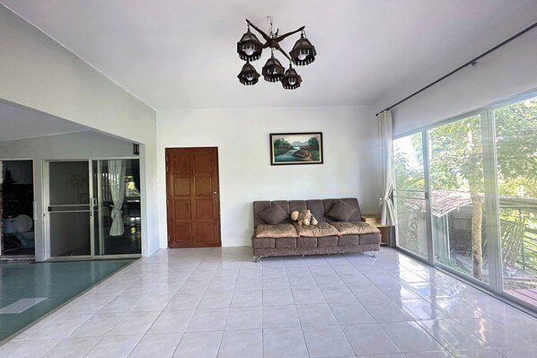A spacious living area with a large window, a sofa with cushions, a wooden door, and a ceiling light fixture.
