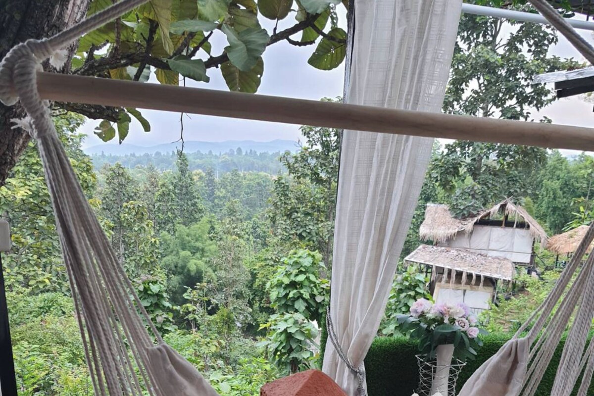 A hammock chair with a cushion, set against a backdrop of lush greenery and a distant view of a traditional house.