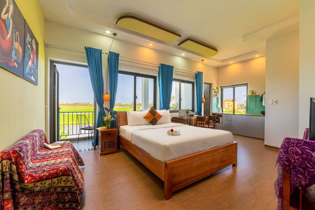 A spacious bedroom with a large bed, a balcony with blue curtains, and a view of a green landscape.