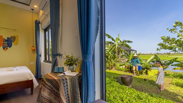 A room with a bed, a table with a patterned tablecloth, and a view of a garden with people working. - Lua House (Da Nang)