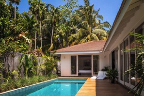 A swimming pool with a wooden deck, surrounded by lush tropical vegetation and a building with large windows. - Good Story Village (Ahangama)