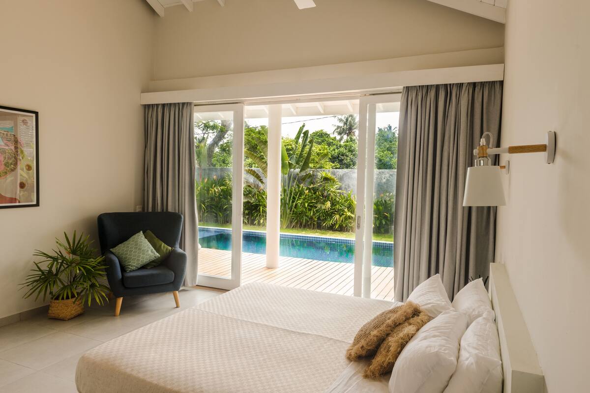 A bedroom with a large bed, a chair, a potted plant, and a view of a swimming pool through glass doors.