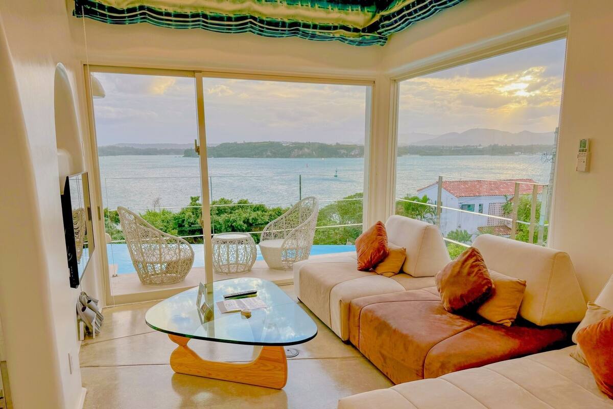 Uma sala de estar moderna com uma mesa de centro de vidro, sofá branco e cadeiras de vime. Janelas amplas oferecem vista para o mar e colinas ao longe.