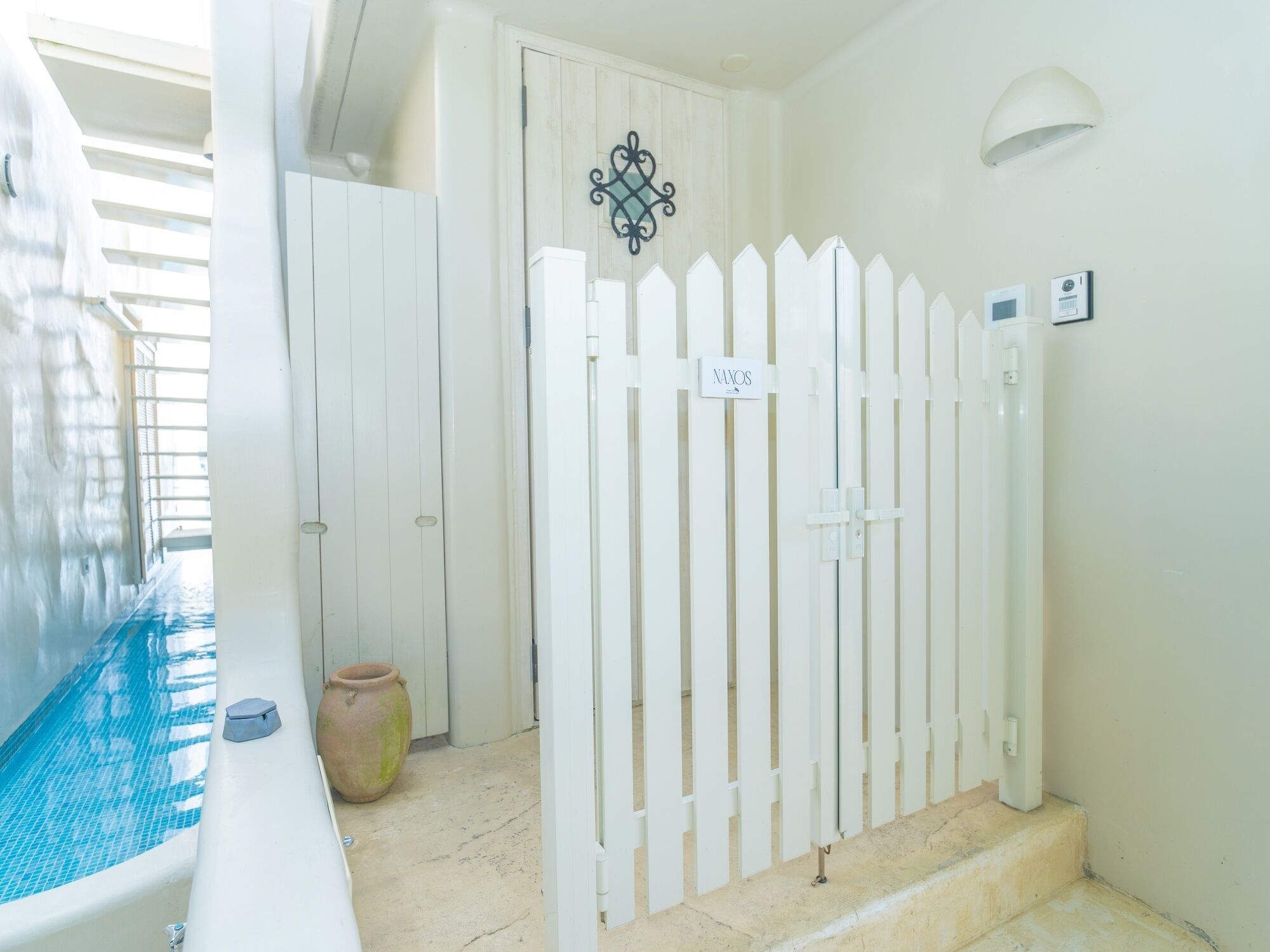 A pool area with a white fence, a door with a 'NAVOS' sign, and a wall-mounted light fixture.