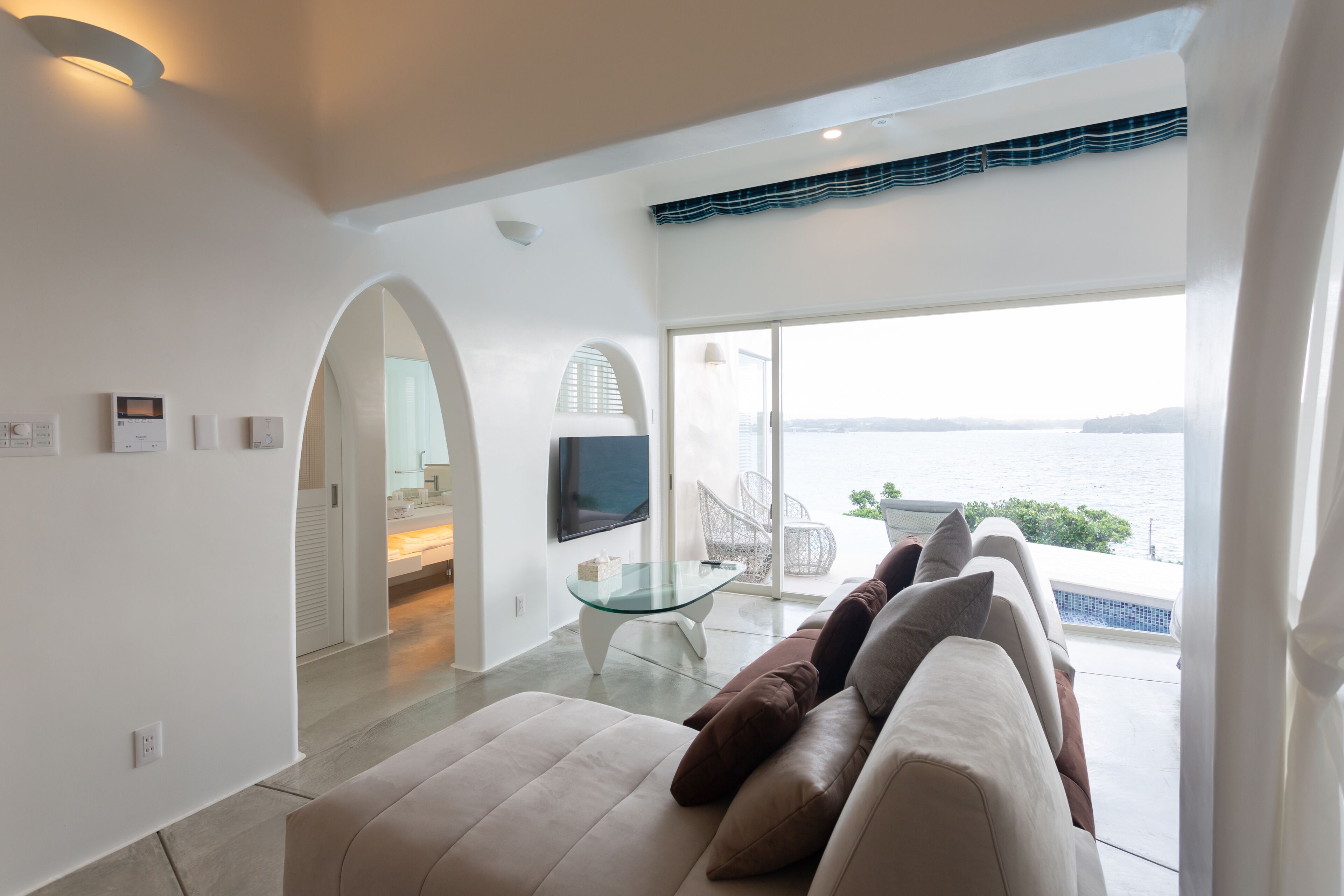 Una sala moderna con un sofá, una mesa de centro de vidrio y un televisor. Se ve el mar a trav és de ventanas grandes.