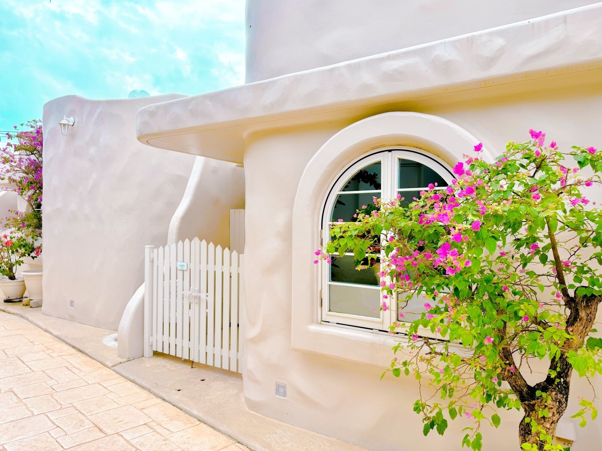 A building with a white facade, a white picket fence, and a window with a view of pink flowers.