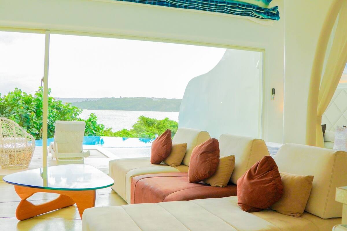 Uma sala de estar moderna com uma grande janela com vista para um corpo de água, uma mesa de centro de vidro e um sofá bege com almofadas.
