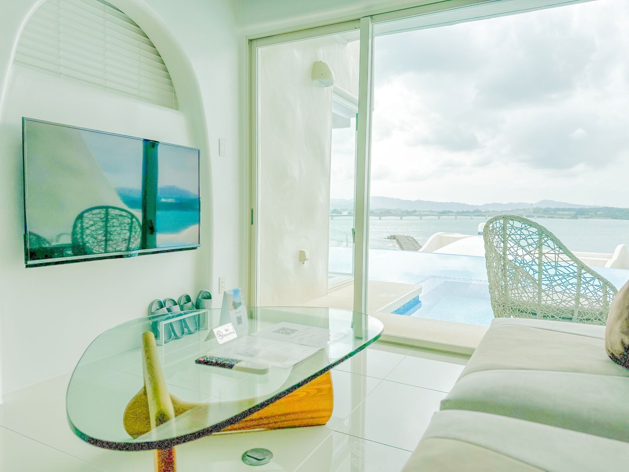 A modern living room with a glass table, a sofa, and a view of a swimming pool and mountains.