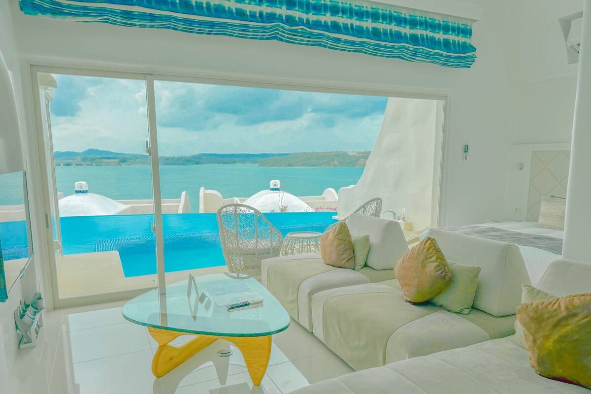 Quarto moderno com mesa de vidro, sofá branco e vista para o mar e montanhas.
