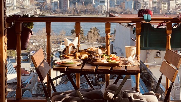 Un balcon avec une table dressée pour un repas, offrant une vue sur un paysage urbain avec des bâtiments et une étendue d’eau.