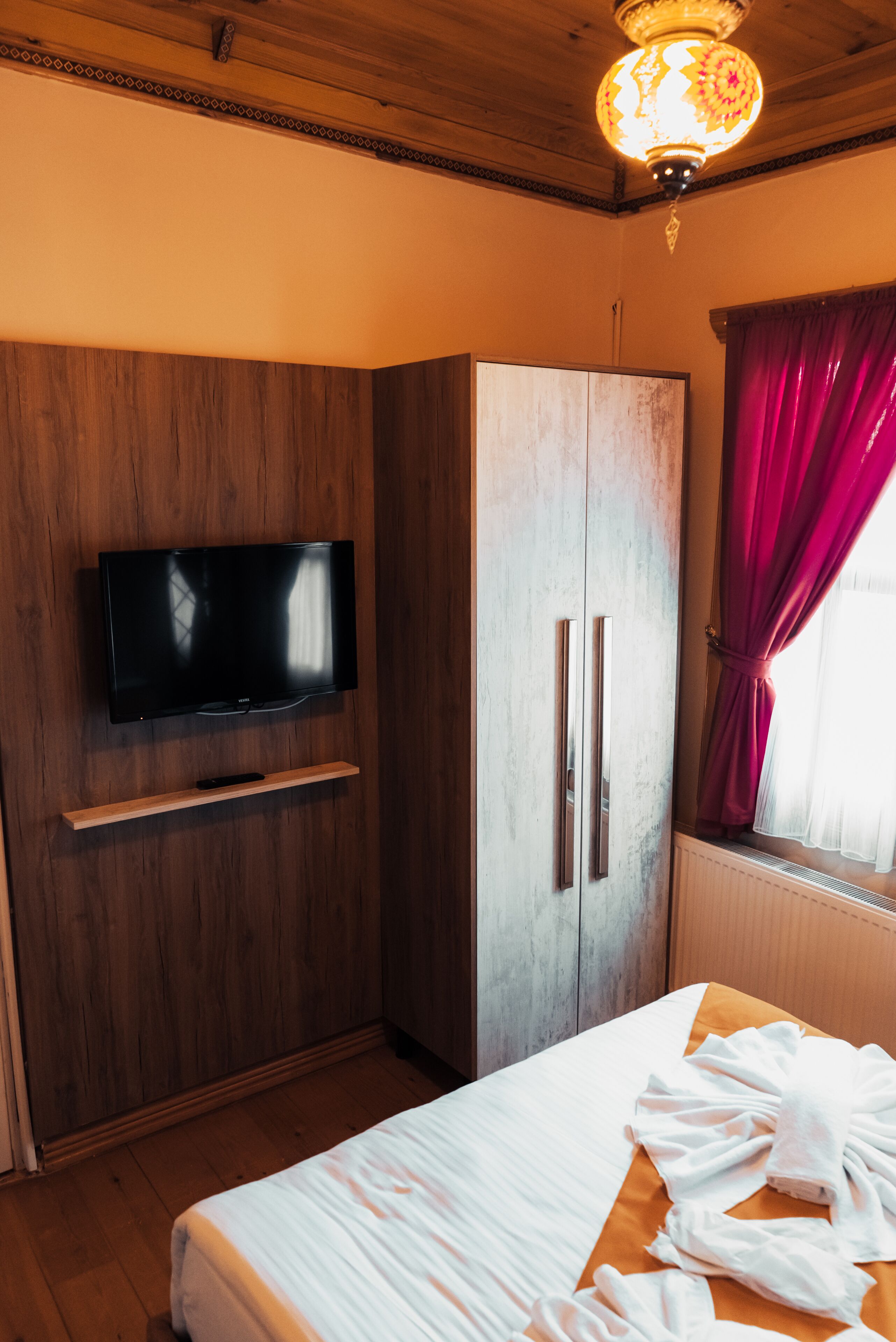 A bedroom with a wooden wardrobe, a flat-screen TV mounted on it, a bed with a white comforter, and a window with red curtains.