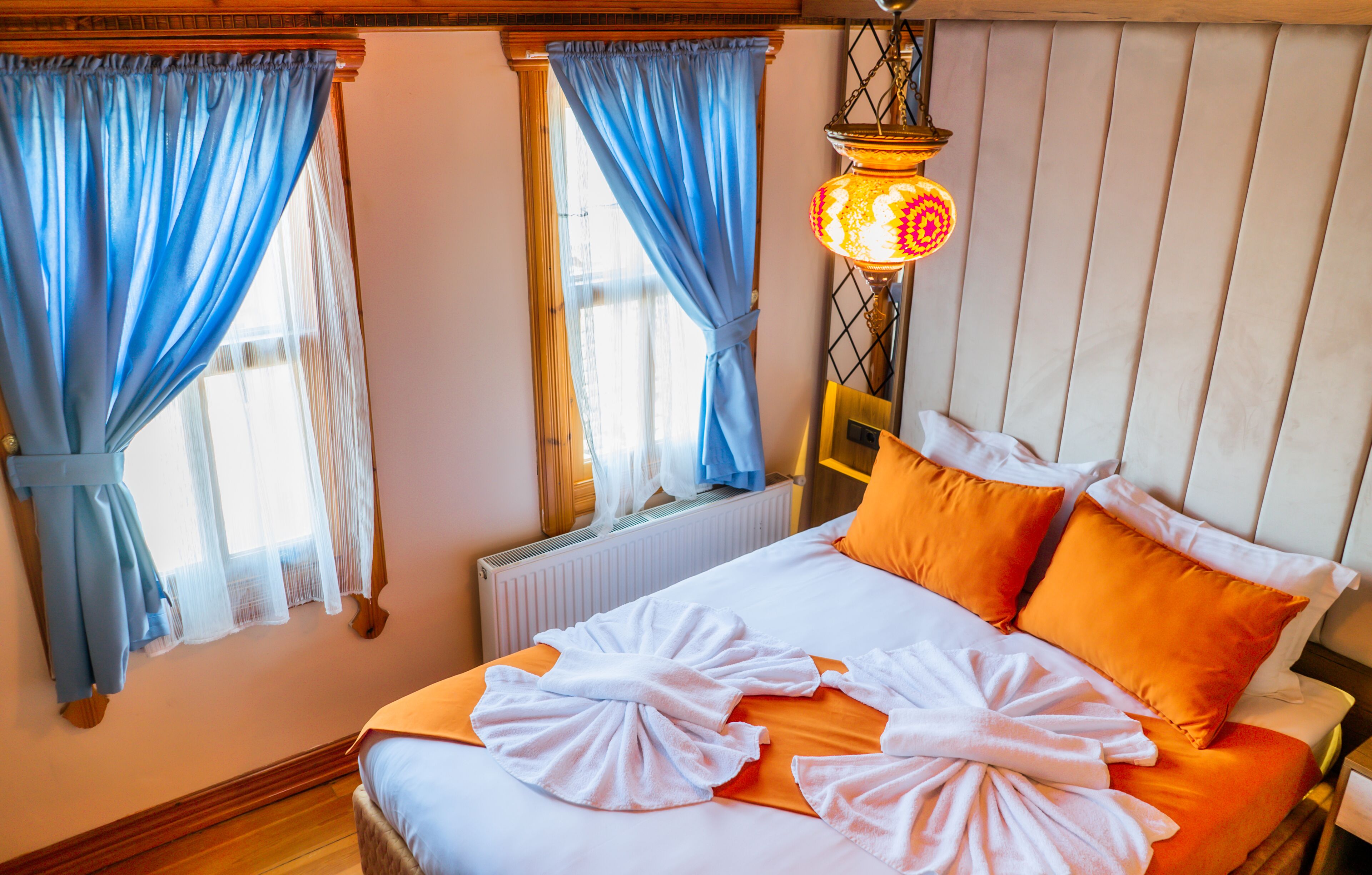 A hotel room with a bed, blue curtains, a radiator, and a hanging lamp.