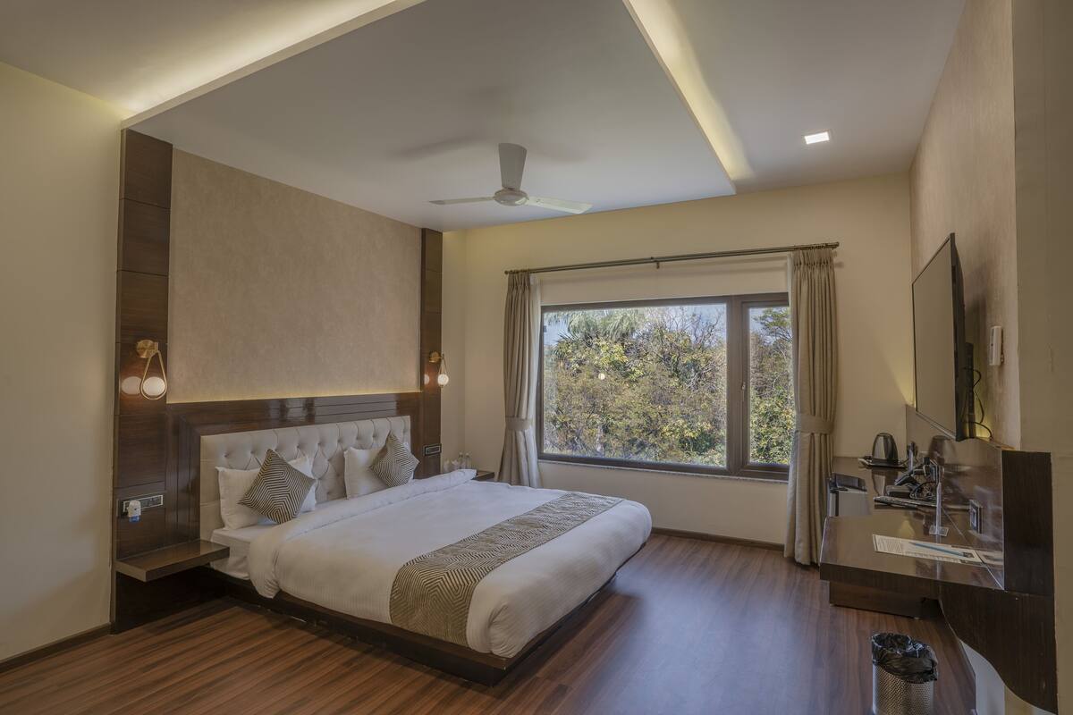 Une chambre d’hôtel moderne dotée d’un grand lit, d’un ventilateur de plafond, d’une fenêtre avec des rideaux, d’une table de chevet avec une lampe, et d’une salle de bain équipée d’un miroir et d’un lavabo.
