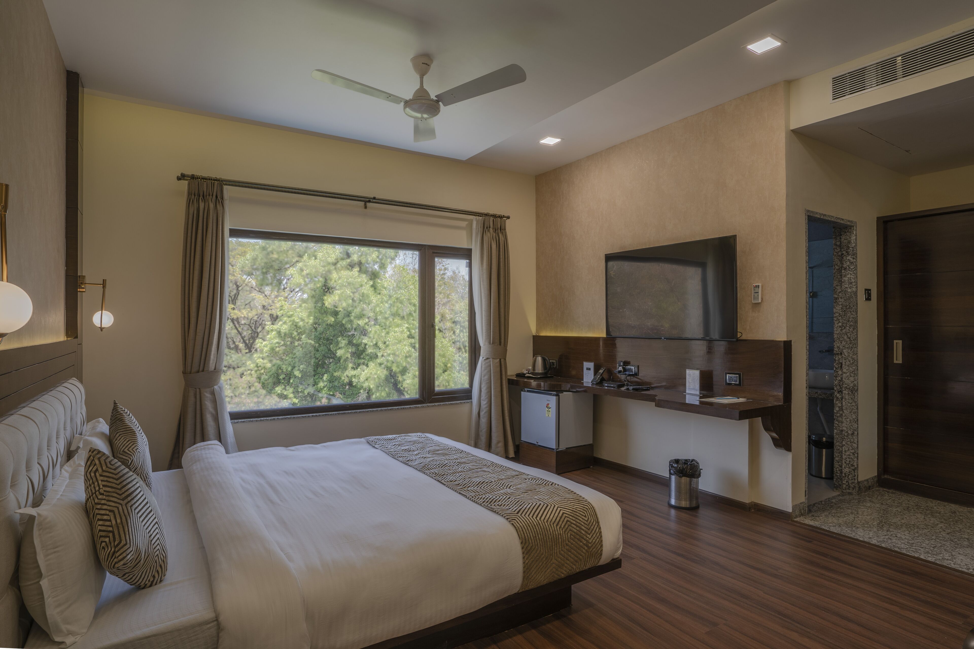 Habitación de hotel con cama, escritorio, espejo, ventilador de techo y ventana con cortinas.