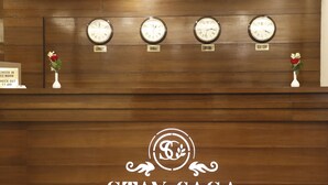 Mostrador de recepción con relojes que muestran la hora en Londres, Dubái, Delhi y Nueva York. Hay dos jarrones con flores a cada lado de los relojes. El mostrador tiene un cartel que indica los horarios de registro.