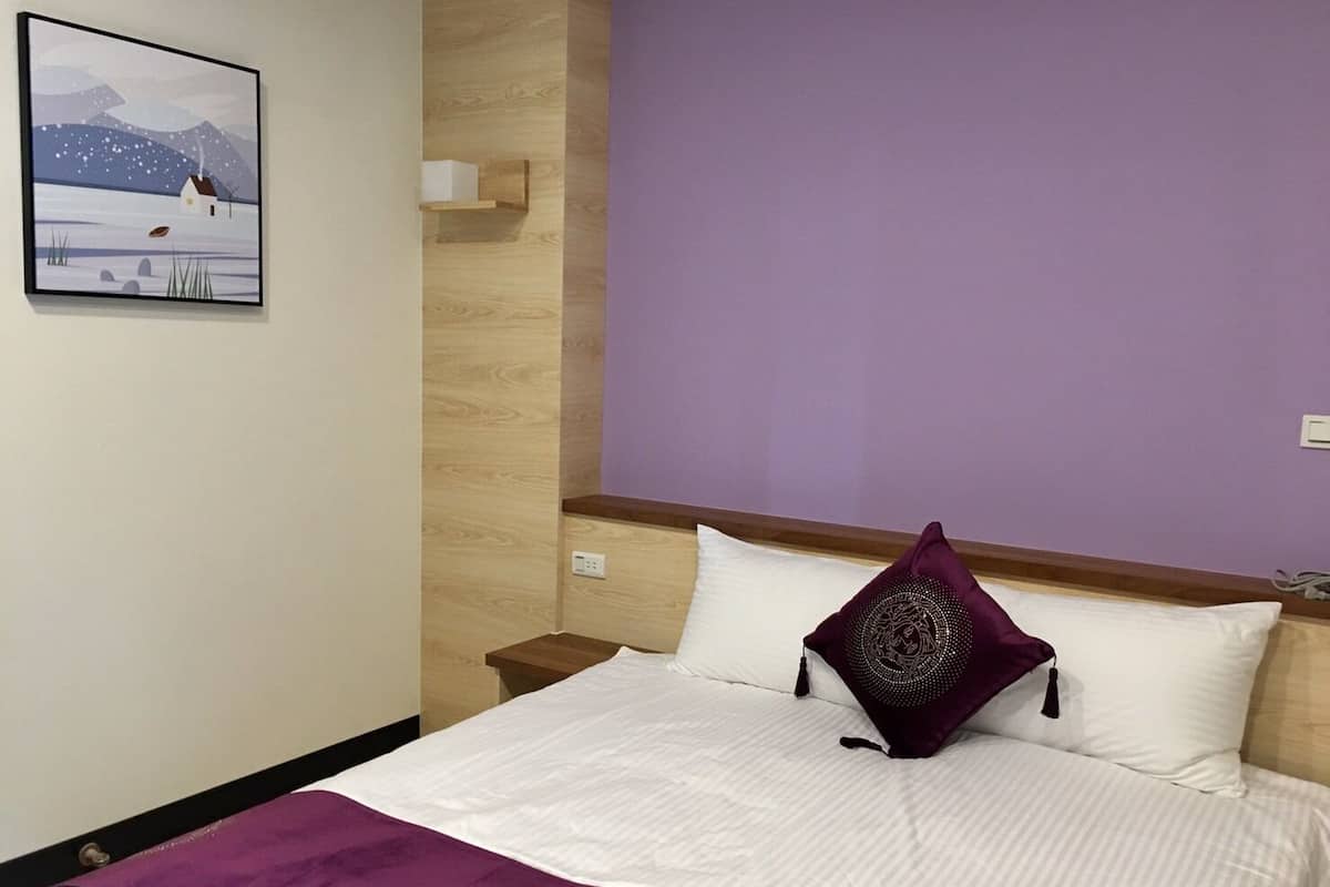 Habitación de hotel con cama, almohada morada, almohada blanca, cabecero de madera, mesita de noche, estante montado en la pared y un cuadro enmarcado con un paisaje nevado.