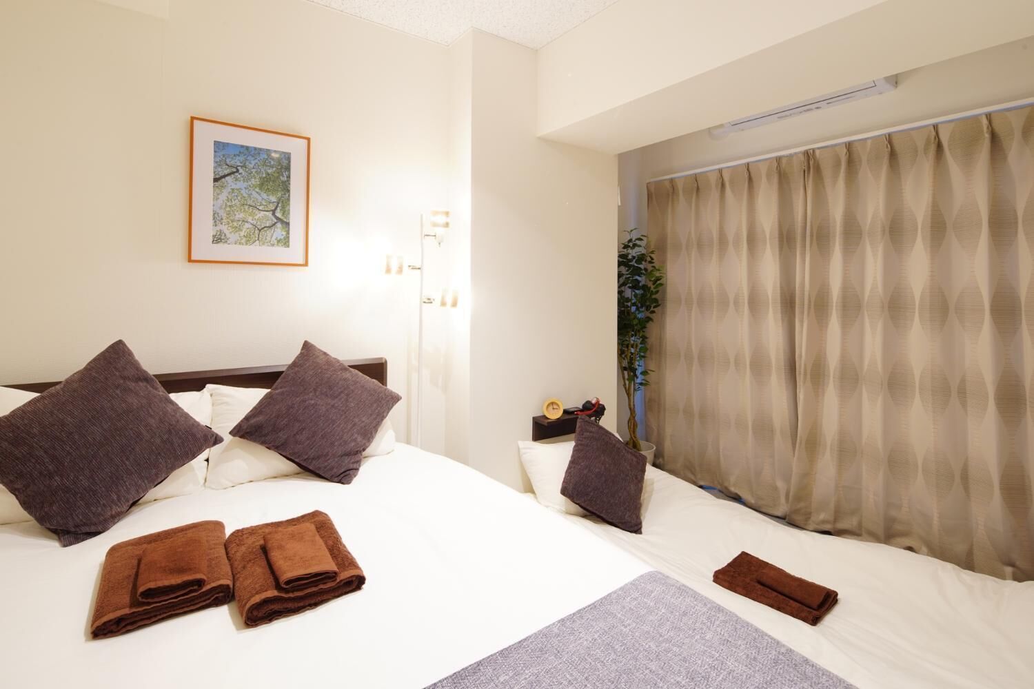 Habitación de hotel con una cama grande, dos almohadas, una mesita de noche con un reloj, una planta y una ventana con cortinas.