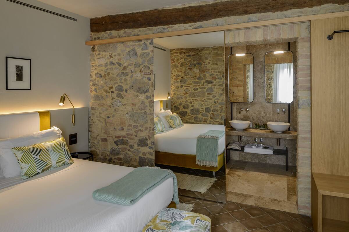 A bedroom with two beds, a stone wall, a bathroom with a sink and mirror, and a wooden ceiling.