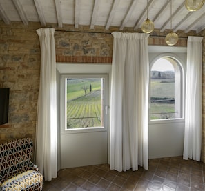 A room with a stone wall, a window with a view of a vineyard, and a patterned chair. - Locanda dei Logi (San Gimignano)