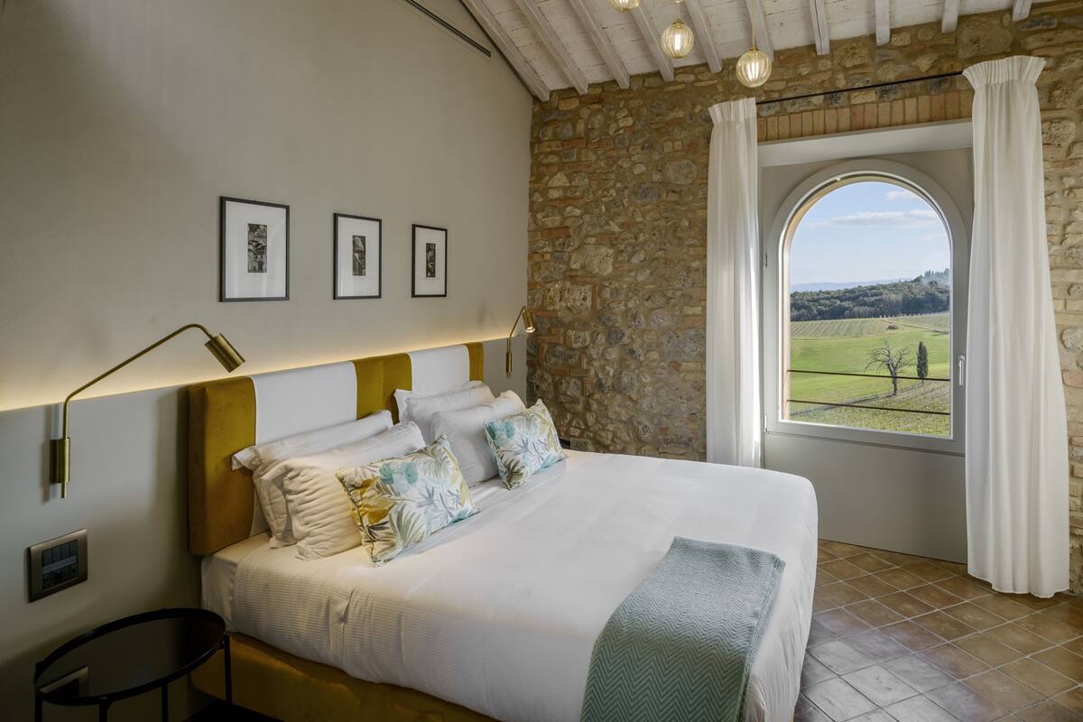 A bedroom with a stone wall, a bed with white bedding, a view of the countryside through a window, and a ceiling with exposed beams and hanging lights.