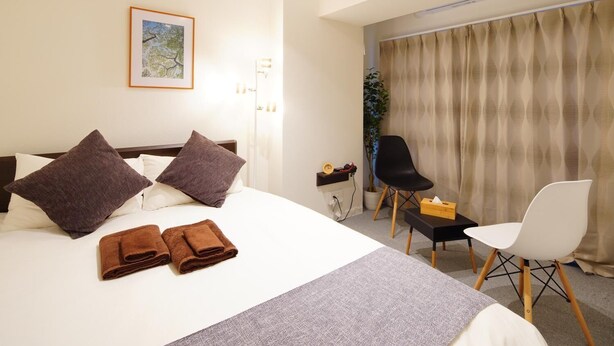 Ett hotellrum med en säng, två kuddar, ett nattduksbord med en klocka, en stol, ett litet bord med en låda och ett fönster med gardiner.